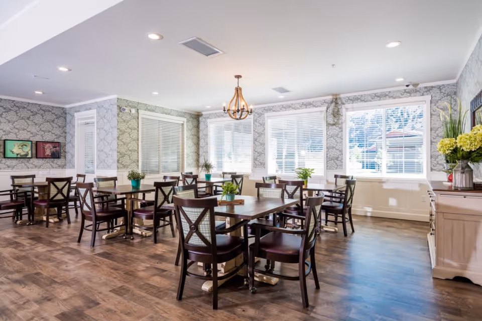 A bright and spacious dining room with multiple wooden tables and chairs arranged neatly. The room features large windows with white blinds allowing natural light to fill the space. There are decorative plants on the tables and a chandelier hanging from the ceiling. The walls have patterned wallpaper and there is a sideboard with flowers and decor on the right side.