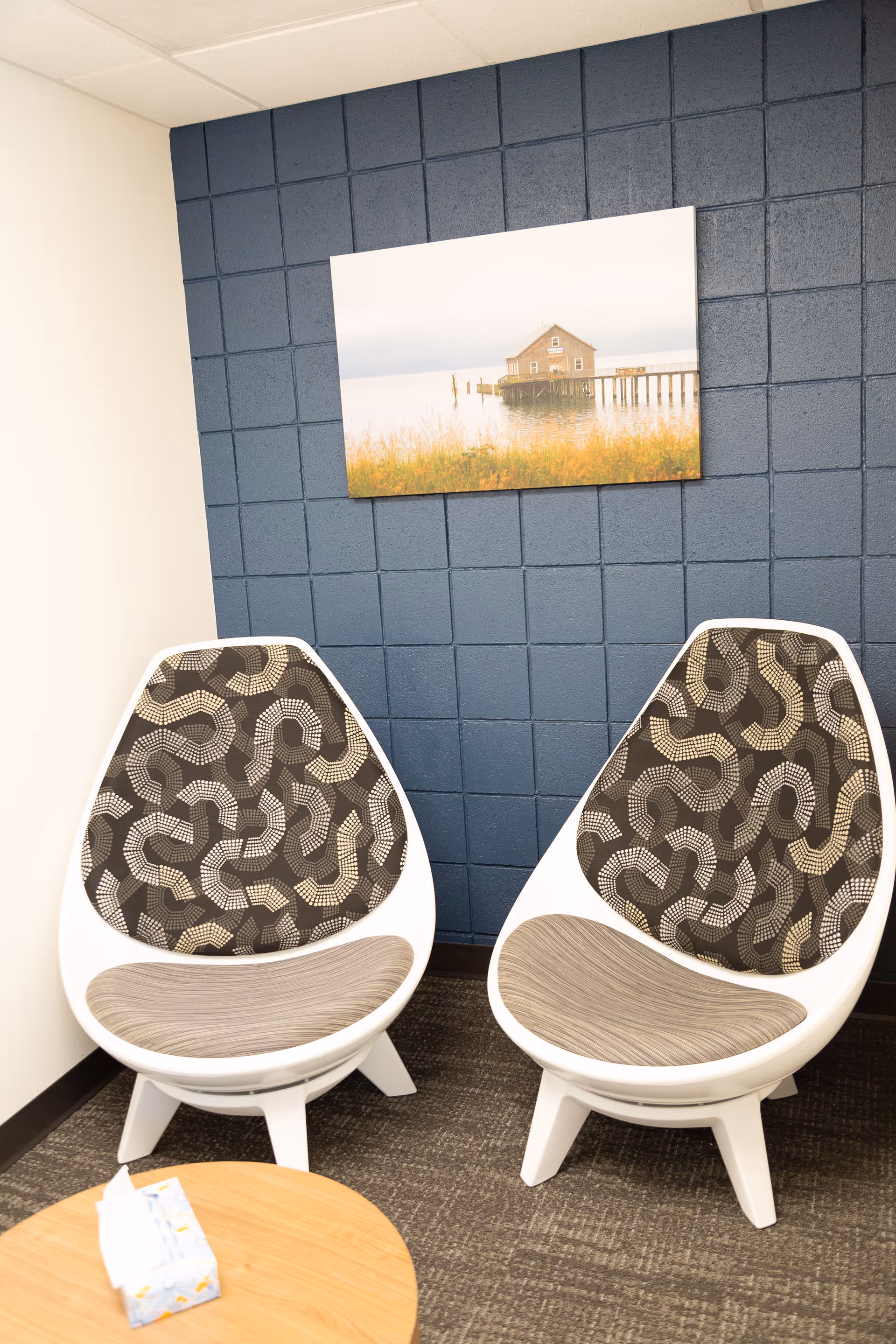 Two modern white chairs with patterned upholstery sit in a blue-tiled lounge with wall art.