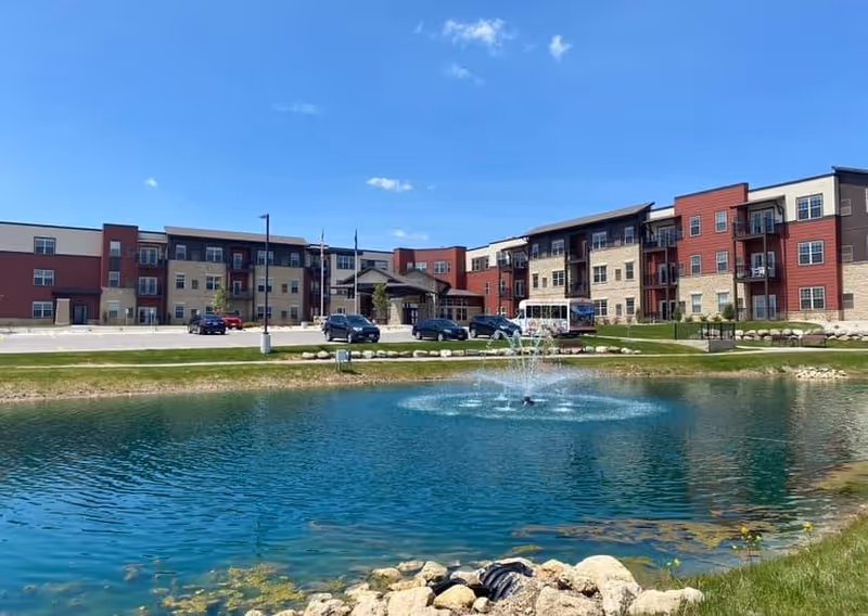 Three-story senior living building with a central entrance, parked cars, and a pond with a fountain in the foreground under a clear blue sky.