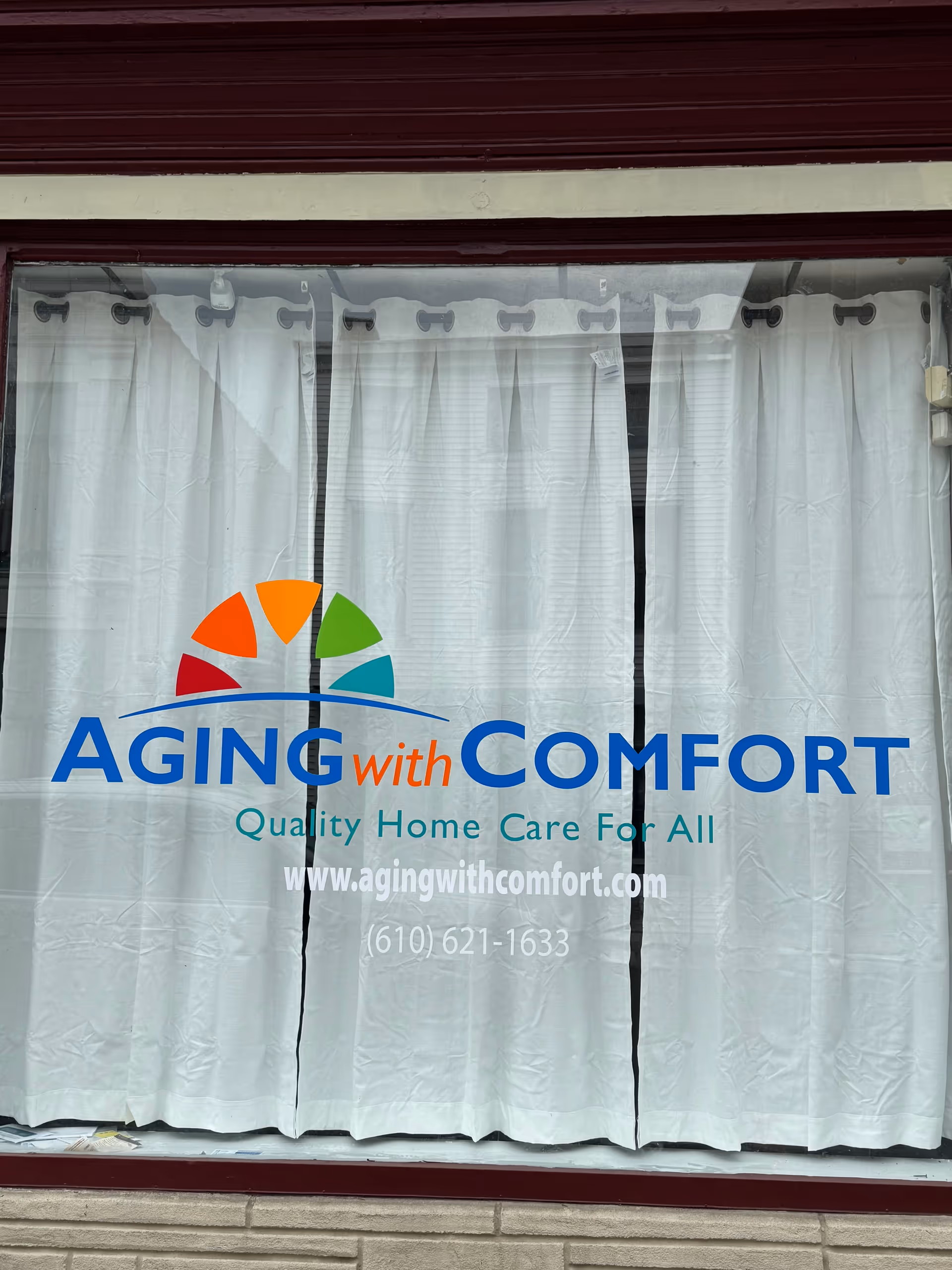 Storefront window with white curtains displaying Aging with Comfort logo and contact information.