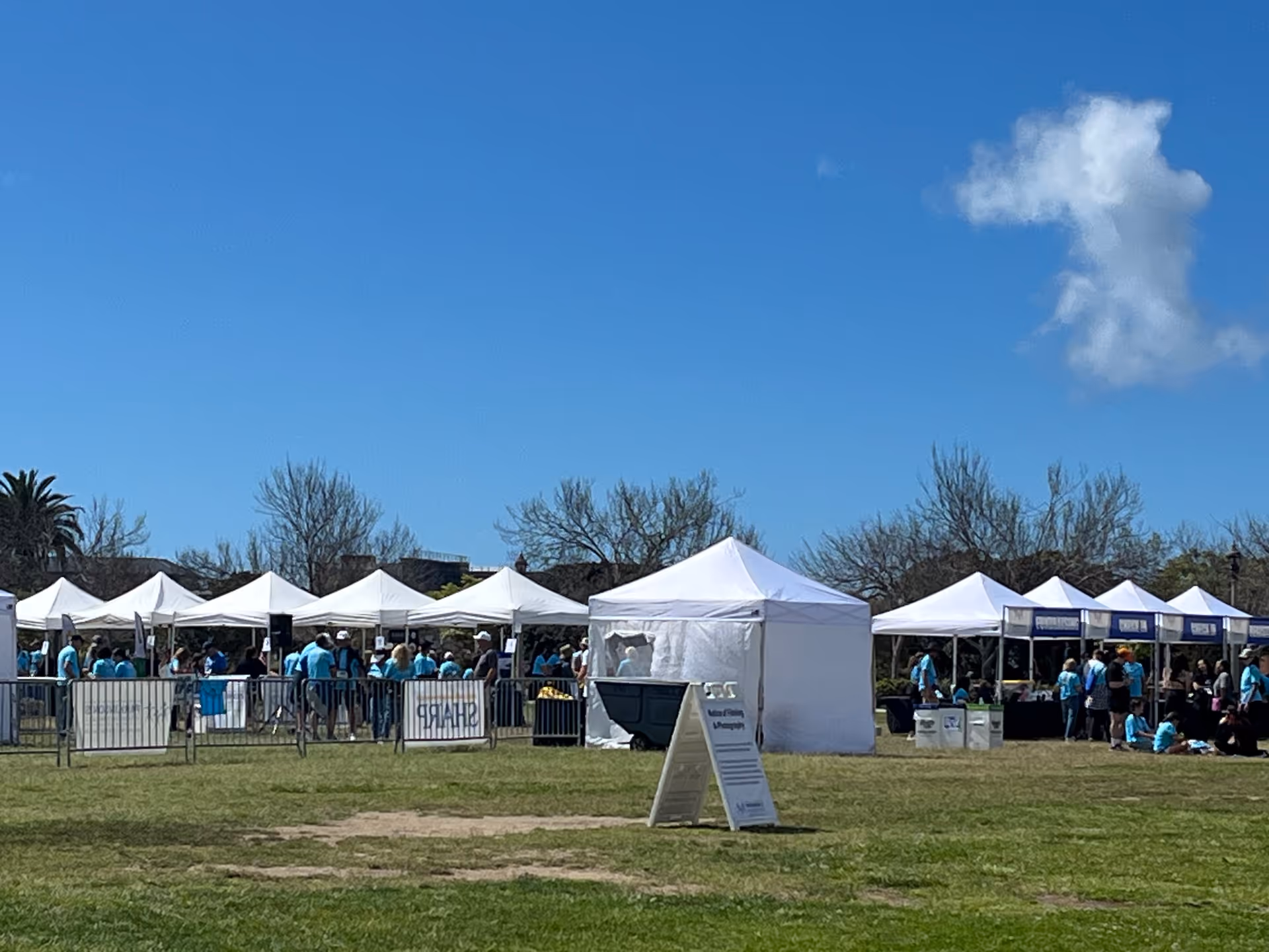 Row of white tents and people gathered for an outdoor event on grass.