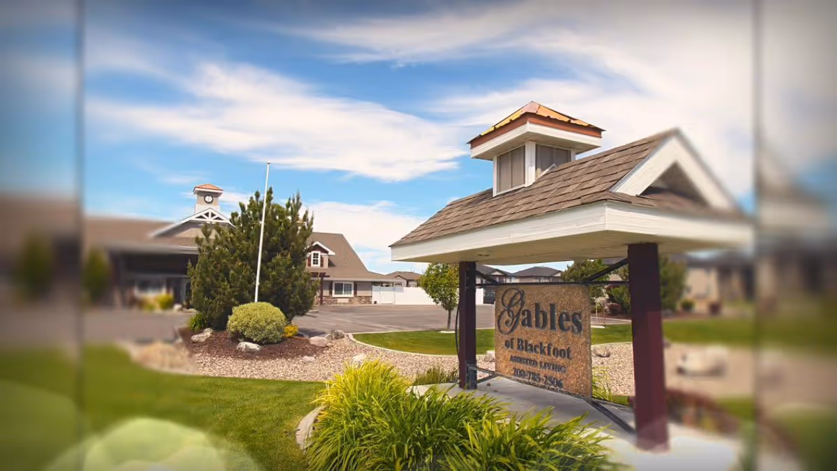 Exterior view of The Gables Memory Care of Blackfoot facility showing a landscaped area with green grass, bushes, and a stone sign under a small roof structure. The building with a peaked roof and clock tower is visible in the background under a partly cloudy blue sky.