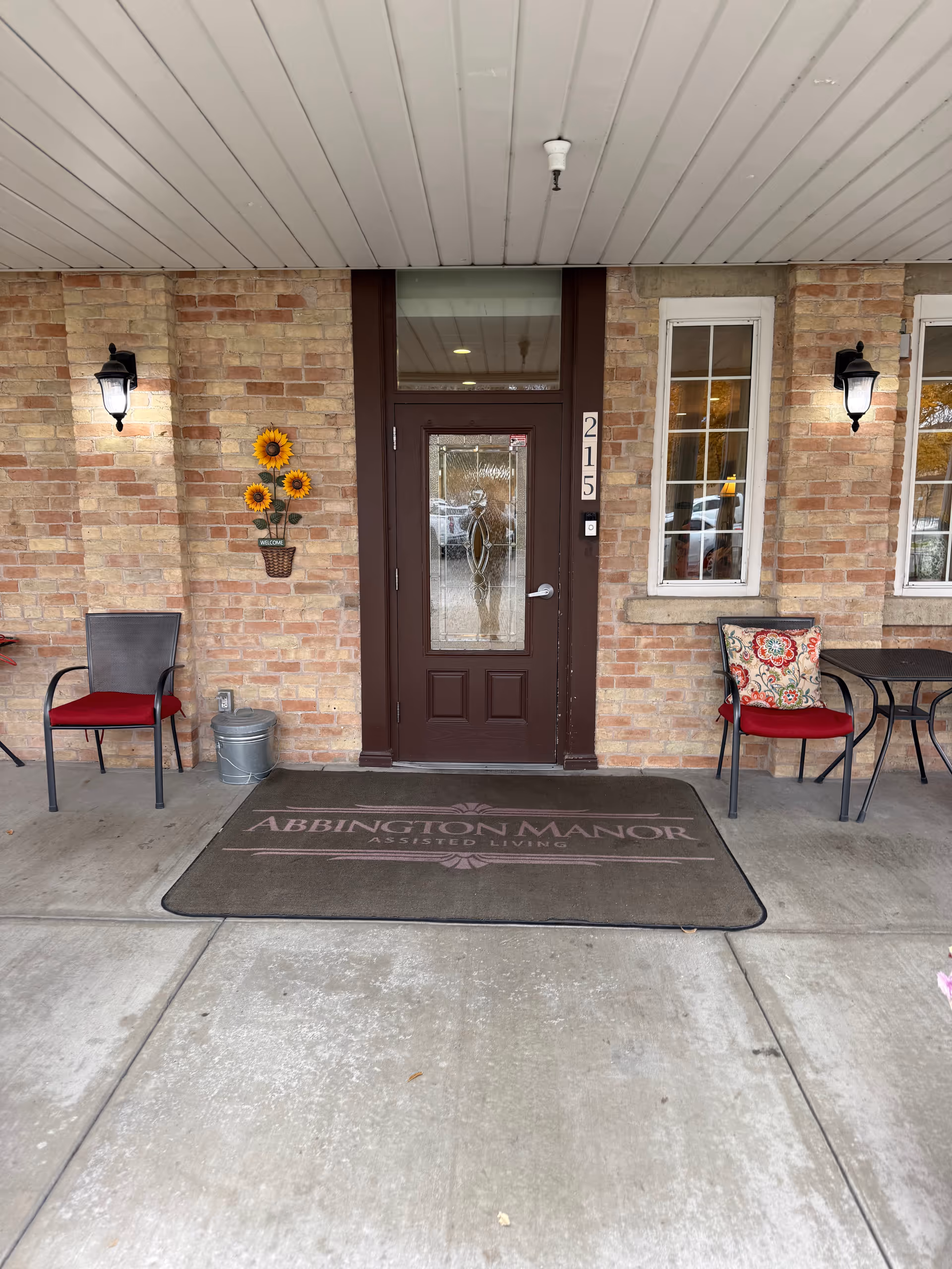 Brick-front entrance of Abbington Manor Assisted Living with seating and sunflowers.