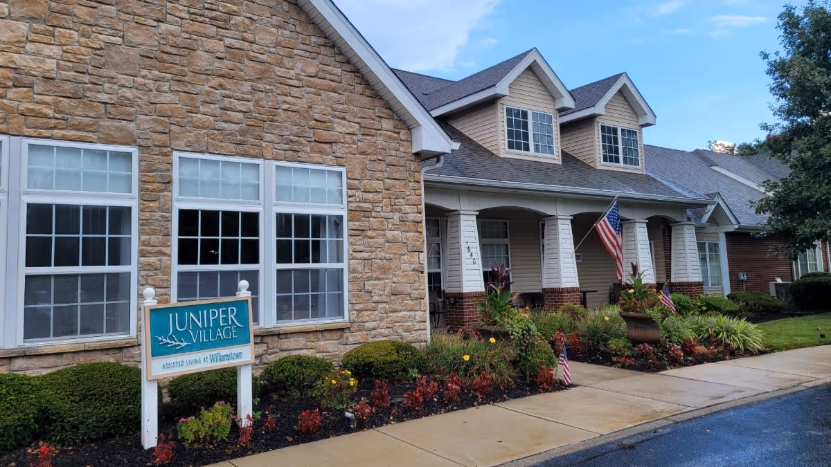 Exterior of Juniper Village at Williamstown with stone facade, entrance, landscaping, and American flags.
