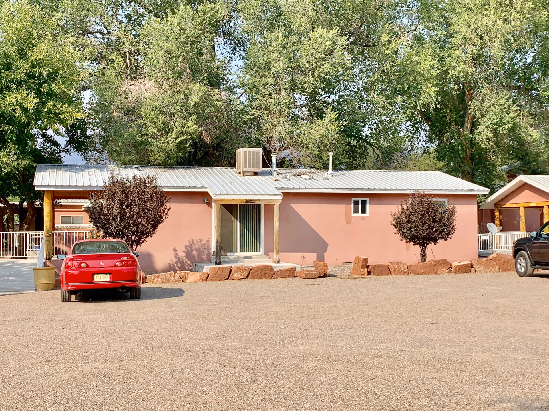 Single-story pink building with a metal roof, two small trees in front, and a gravel parking area with a red car and a black vehicle parked. Large green trees are visible behind the building.