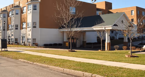 Front exterior of a multi-story assisted living building with a covered entryway, lawn, and sidewalk.