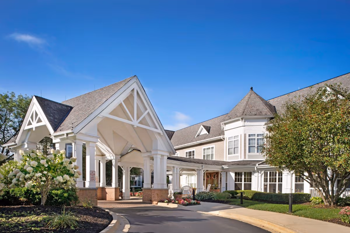 Exterior view of Sunrise of Crystal Lake senior living facility showing a large covered entrance with white pillars and a peaked roof, surrounded by landscaped greenery and flowering bushes under a clear blue sky.