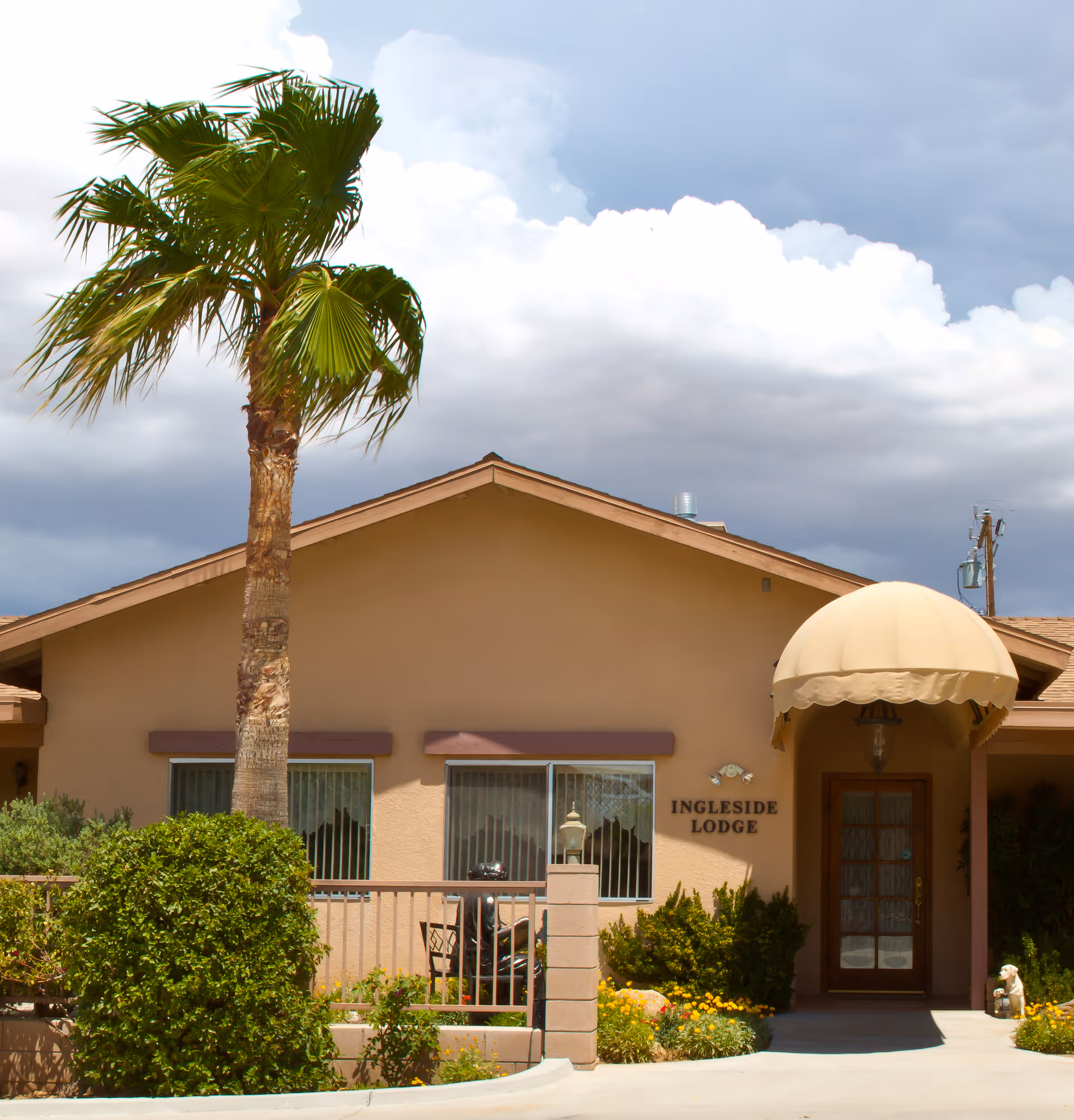 Exterior view of Ingleside Lodge Assisted Living facility showing a single-story building with a beige facade, a palm tree, bushes, flowers, and a covered entrance with an awning.