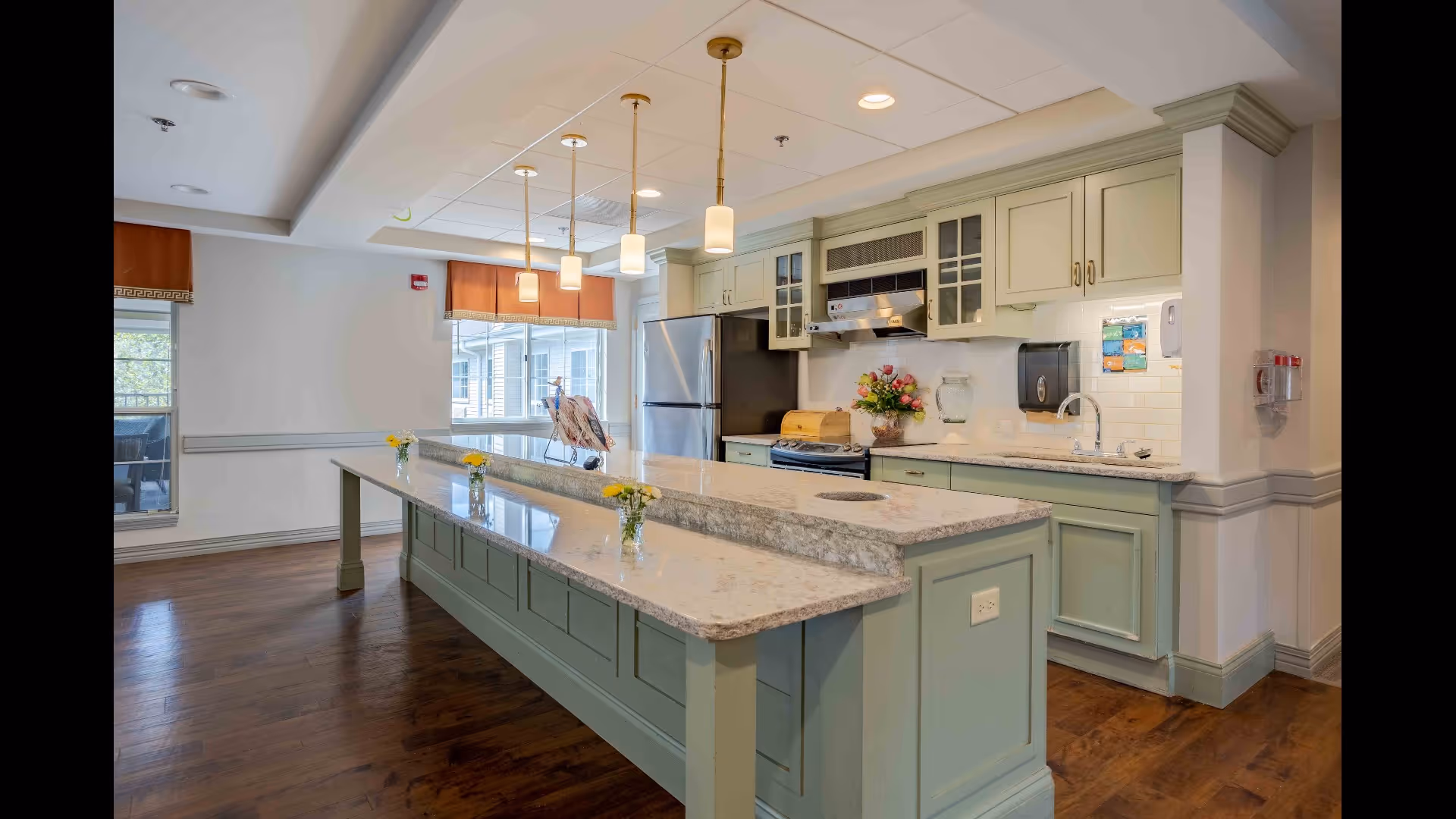 A spacious kitchen area with a large marble countertop island in the center, light green cabinetry, stainless steel refrigerator, stove, and a sink. The kitchen is well-lit with pendant lights hanging above the island and natural light coming through the windows with orange valances. There are small vases with yellow flowers on the island and a bouquet of flowers near the stove.