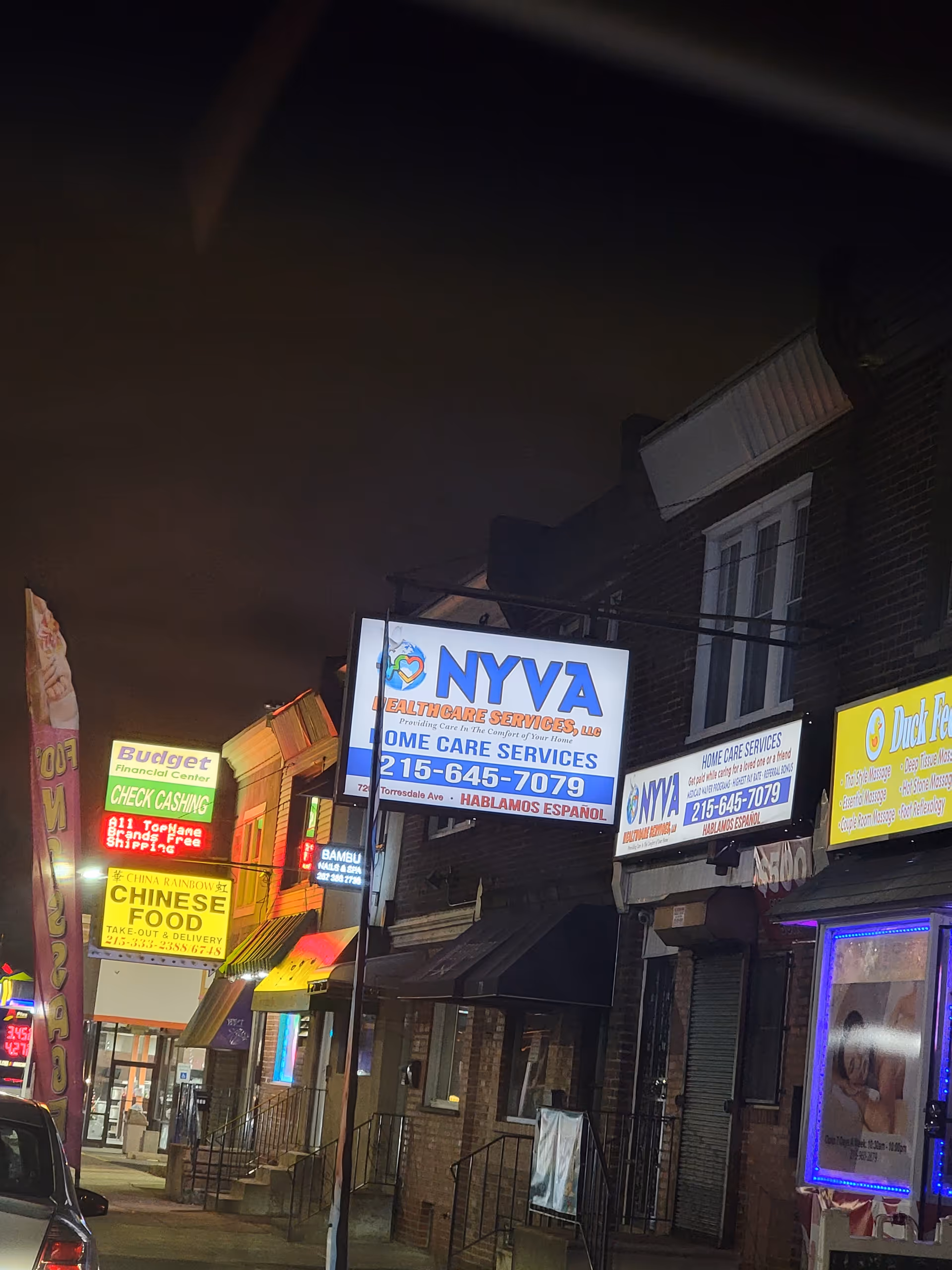 Nighttime street view with NYVA Healthcare Services sign among illuminated storefronts