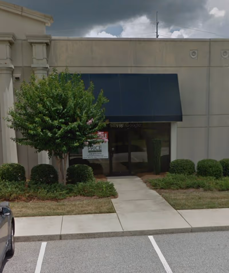 Front of a beige building with a black awning, glass entrance, and manicured shrubs.