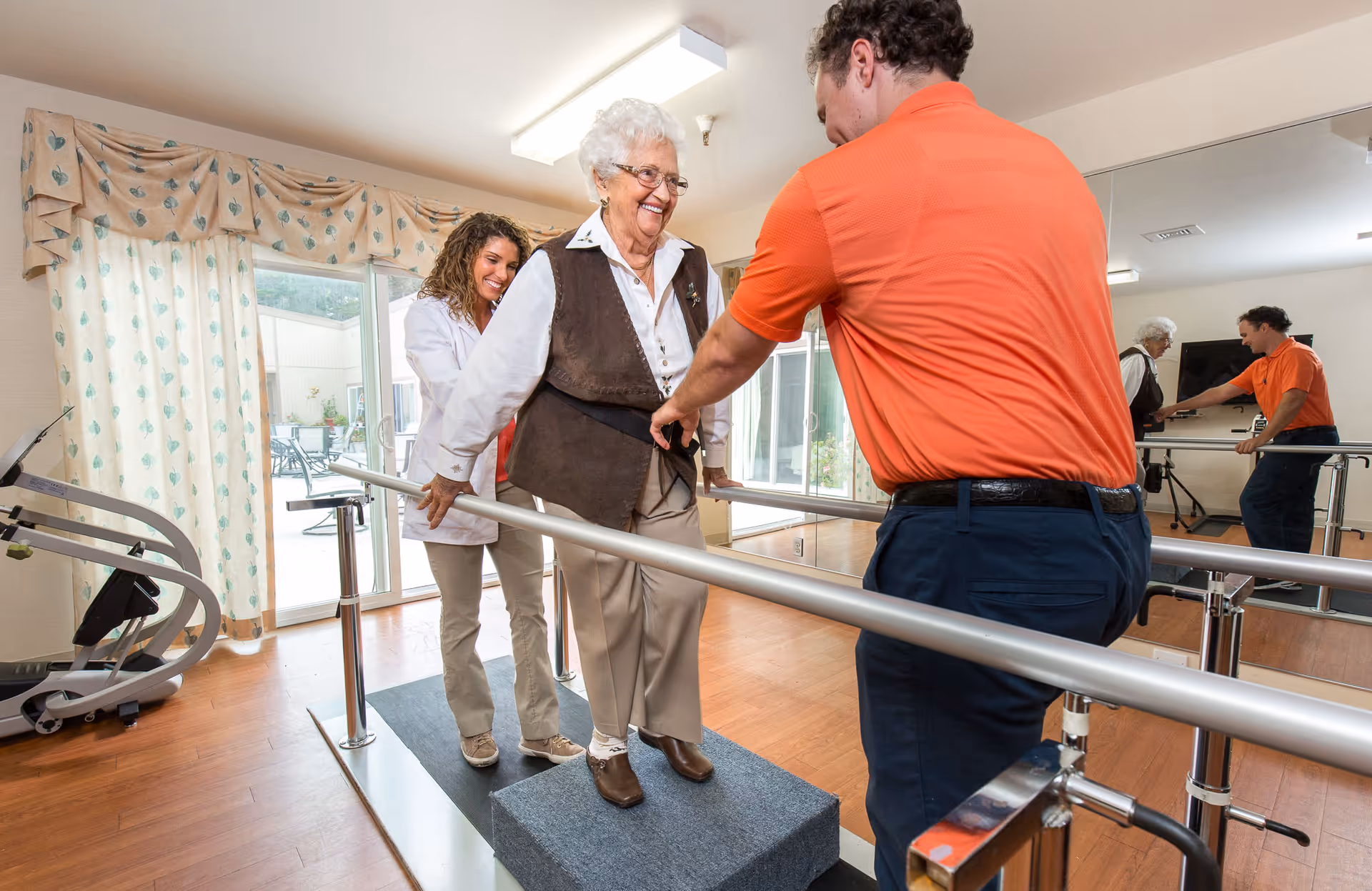 An elderly woman is walking on a raised platform between parallel bars in a rehabilitation or physical therapy room, assisted by a female healthcare professional and a male therapist wearing an orange shirt. The room has wooden floors, large windows with curtains, and a mirrored wall reflecting the scene.
