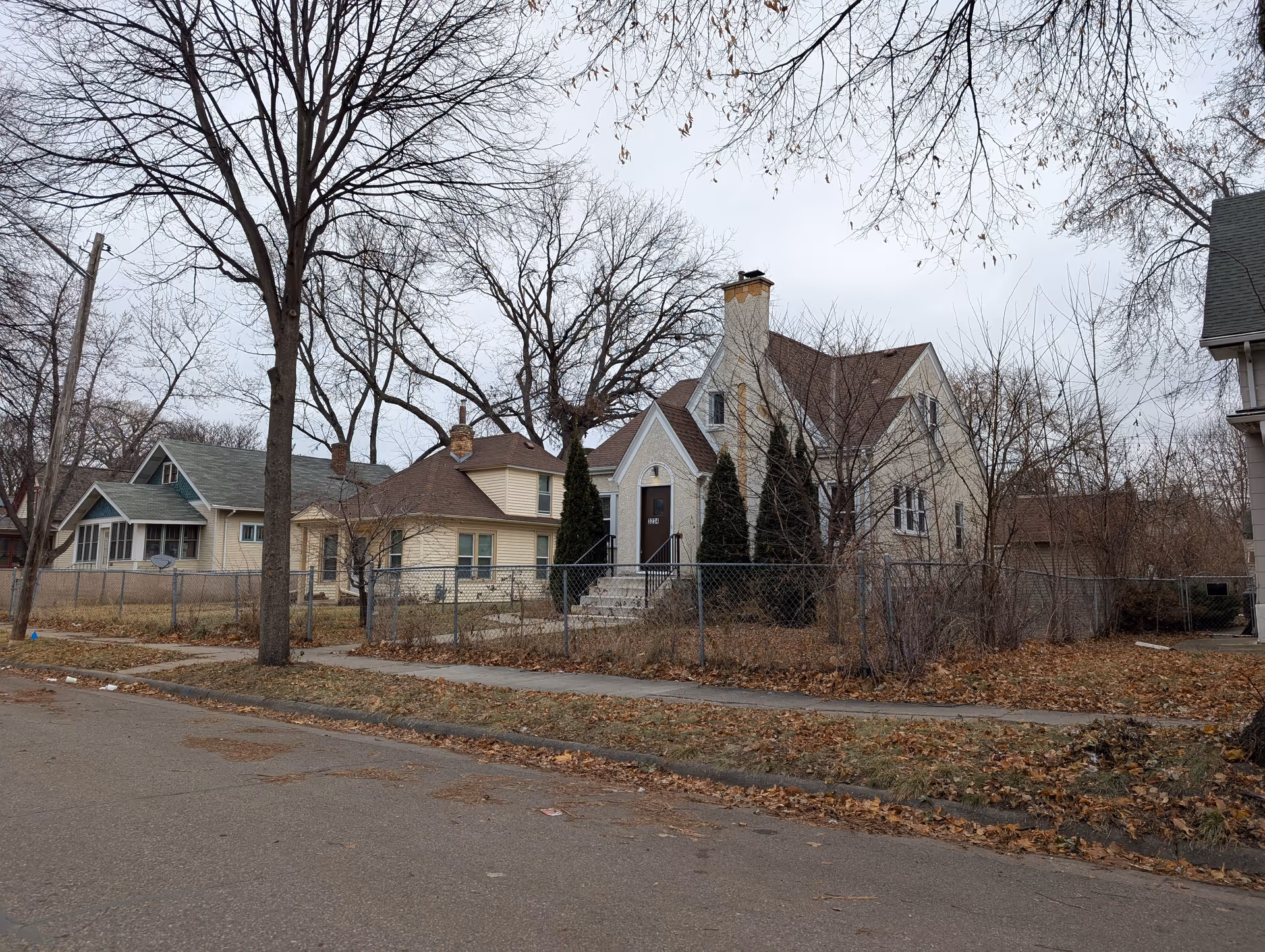 Cream-colored two-story house with a front chimney, fenced yard, and leafless trees along a quiet residential street.
