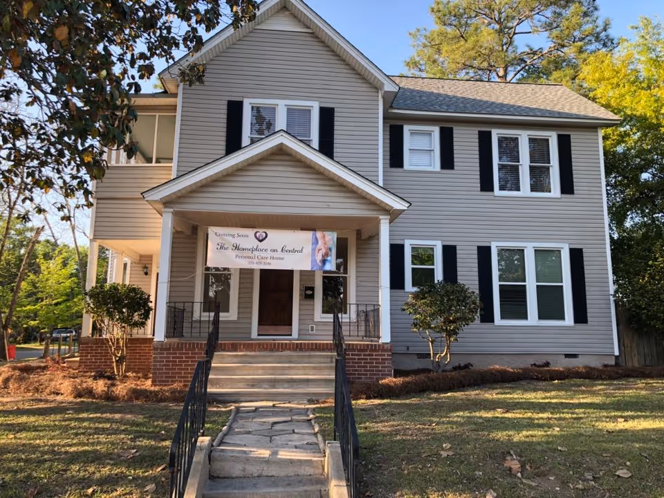 Two-story beige-gray house with black shutters and a banner for The Homeplace on Central.