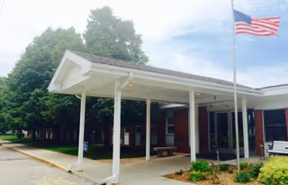 Covered entrance/portico of a brick nursing home with benches, landscaping, and an American flag on a flagpole.