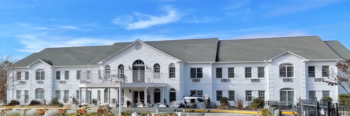 Exterior view of a large two-story white brick building with multiple windows and a central balcony, under a blue sky with some clouds. The building is surrounded by a landscaped area with plants and a parking lot in front.