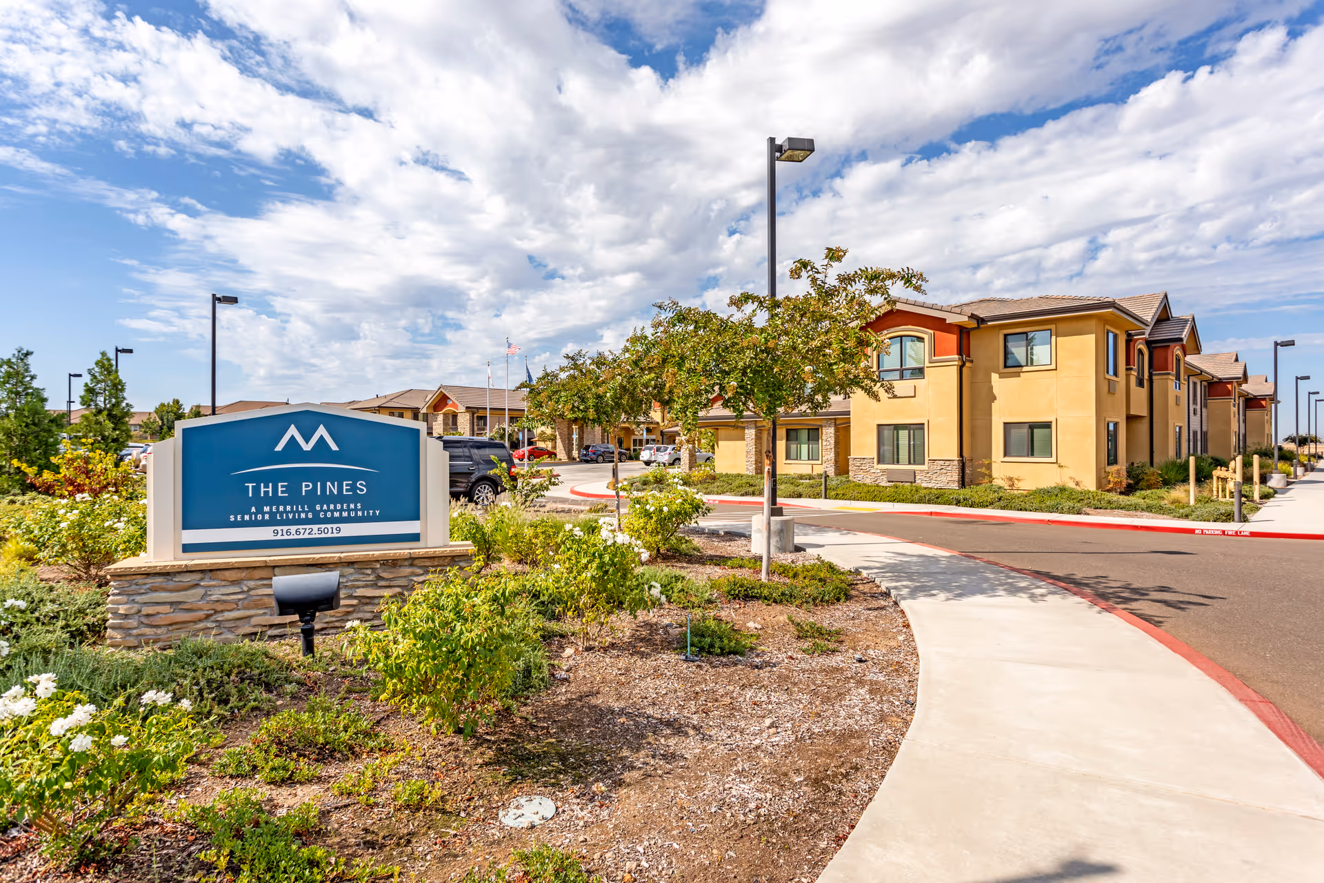 Exterior view of The Pines, A Merrill Gardens Community, with sign, curved sidewalk, and tan buildings.