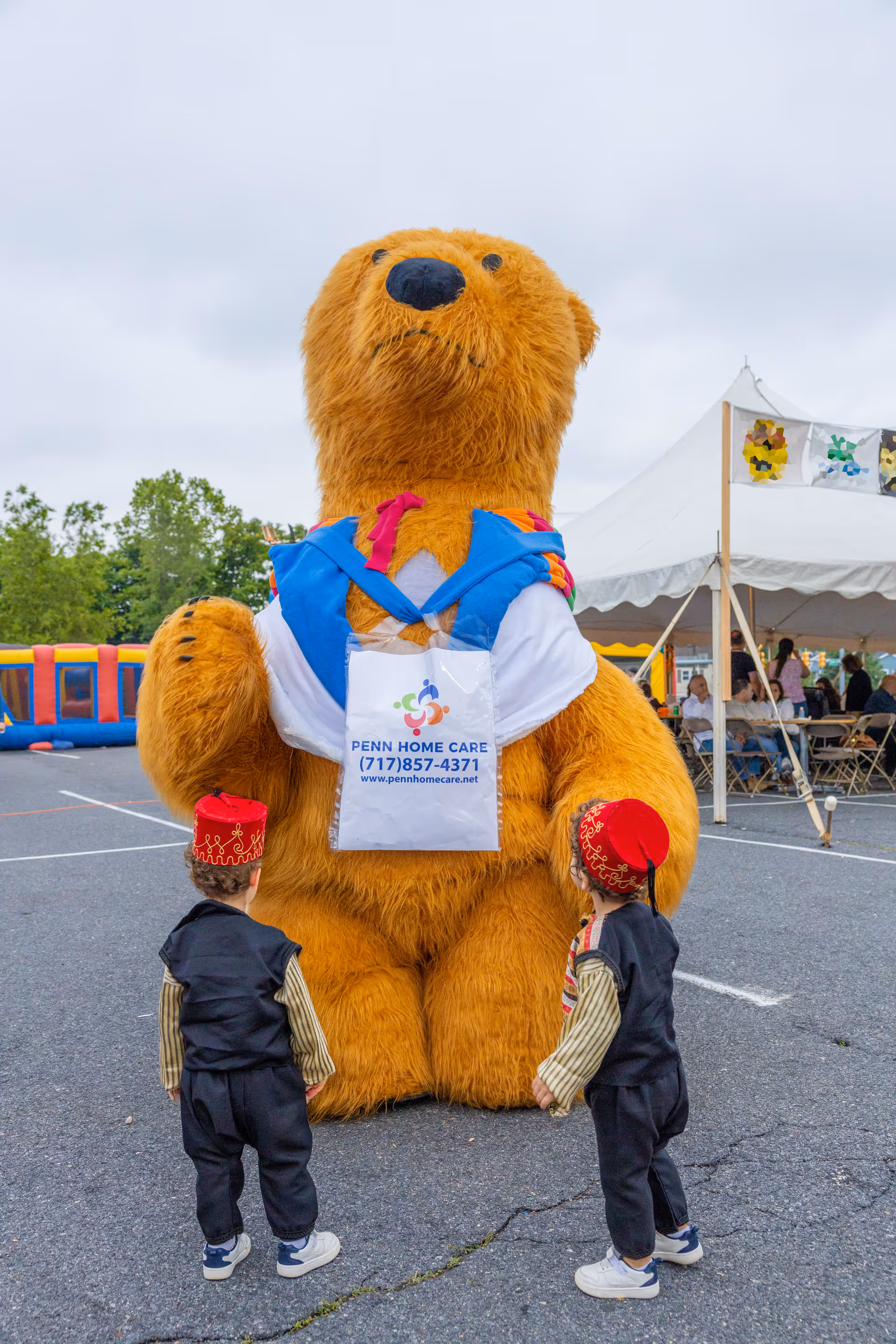 Large brown bear mascot wearing a Penn Home Care tote stands outdoors with two children nearby.