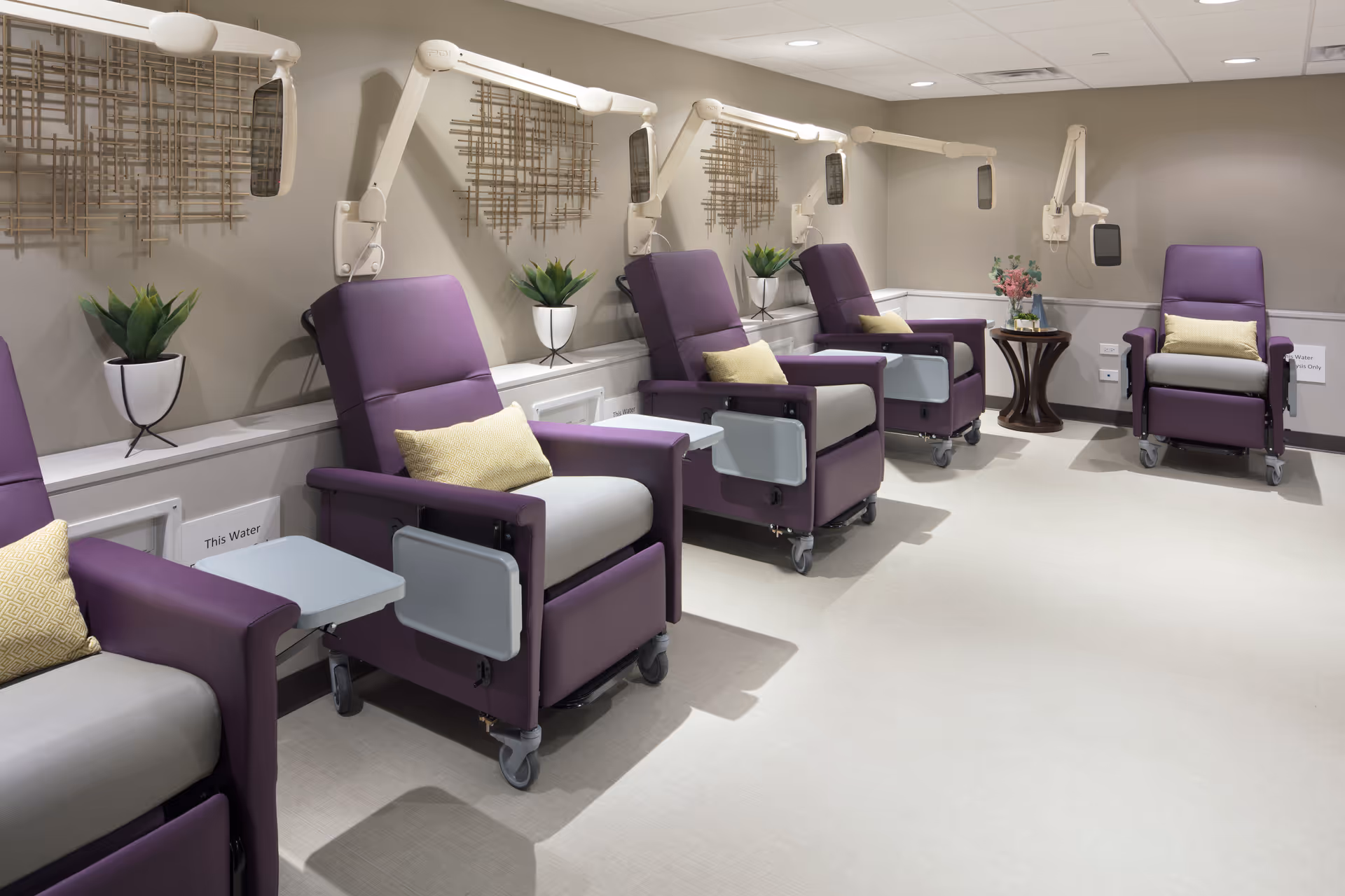 Row of purple reclining chairs with attached trays in a clean, well-lit medical room.