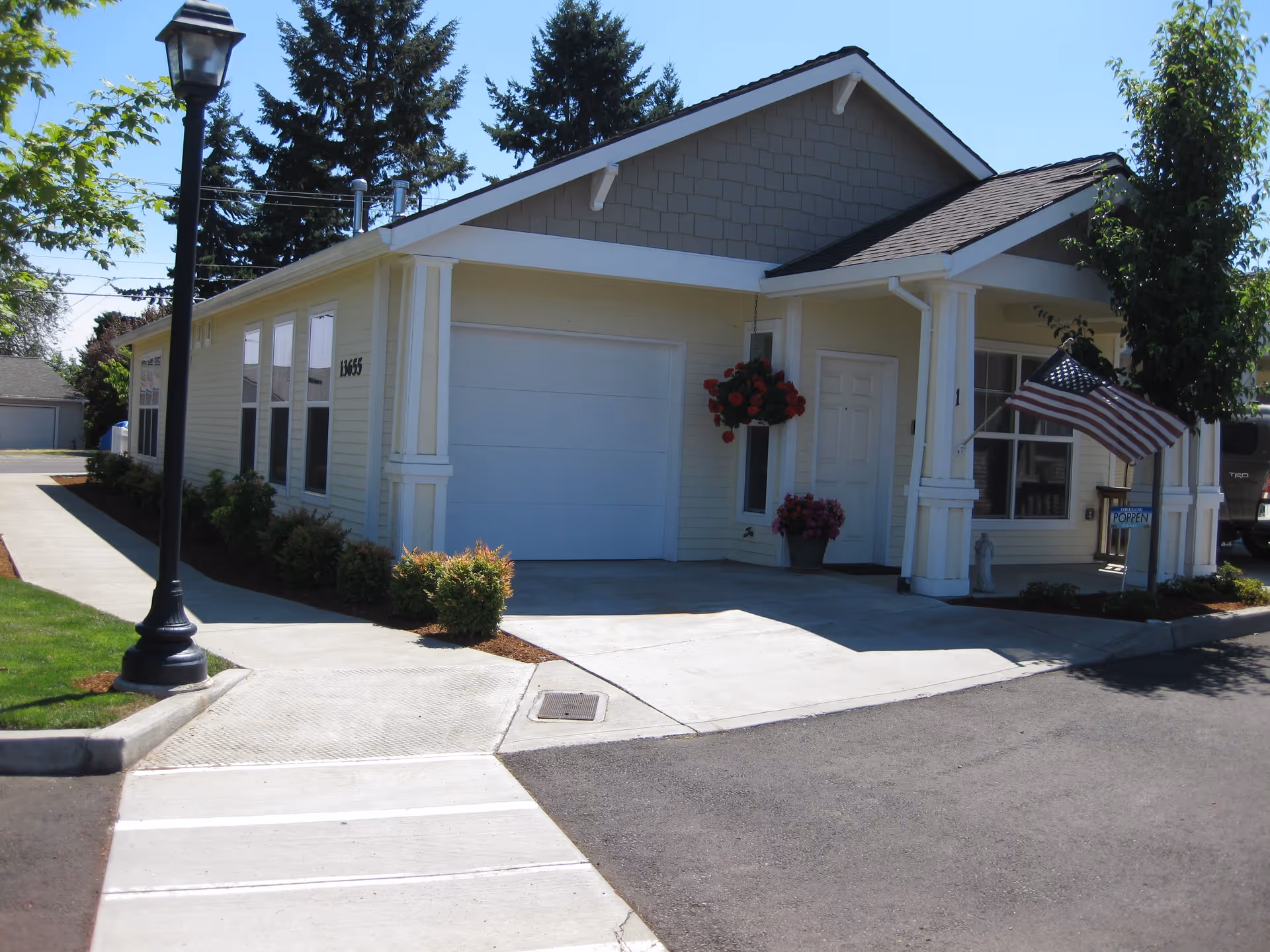 Exterior view of a single-story building with light yellow siding and a dark shingled roof. There is a garage door, a white entrance door, and several windows. The building is surrounded by a sidewalk, small bushes, a lamp post, and a tree. An American flag is displayed near the entrance, and a sign indicating 'OPEN' is visible.