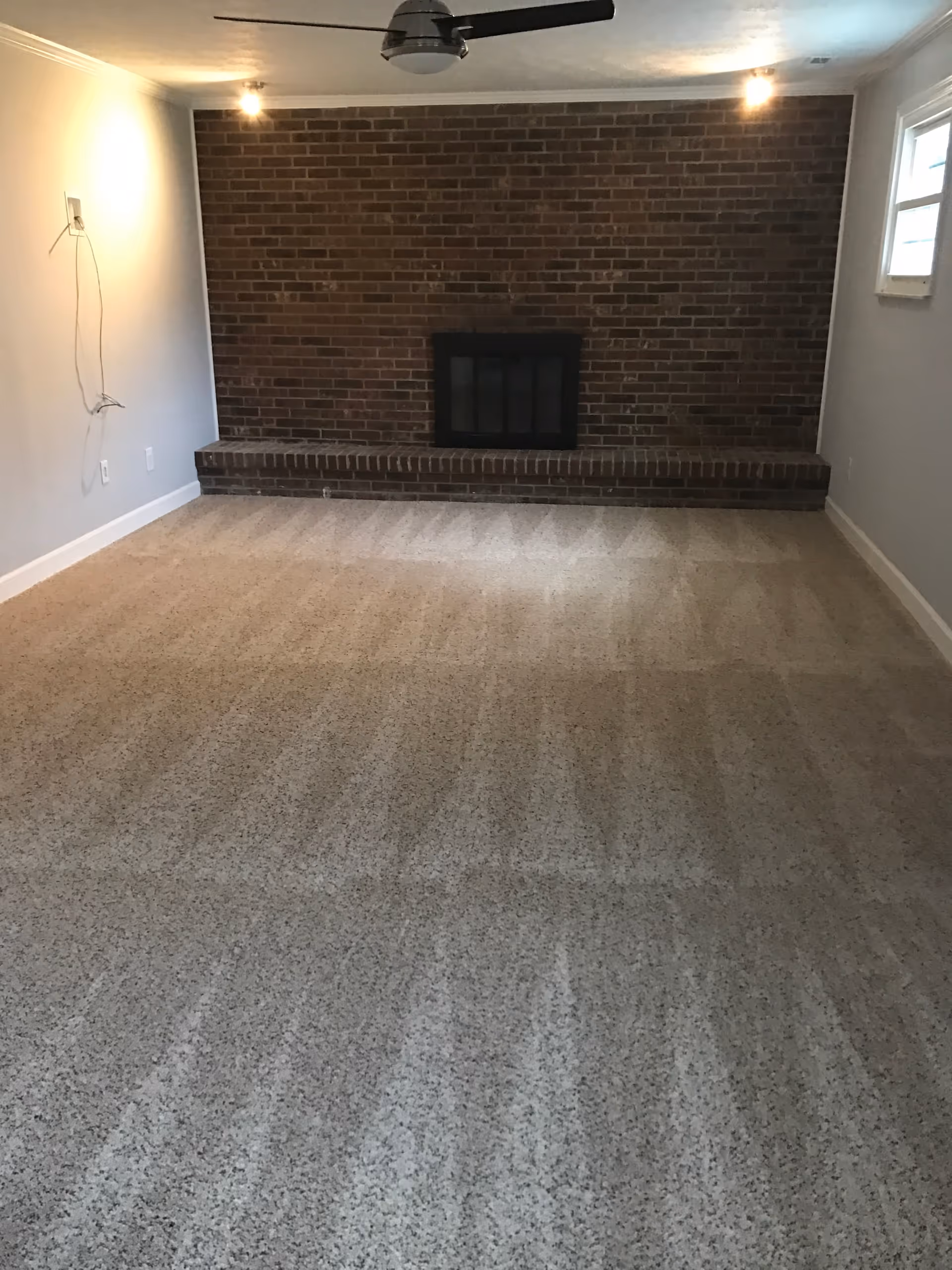 Empty living room with brick fireplace, carpet, ceiling fan, and a small window.