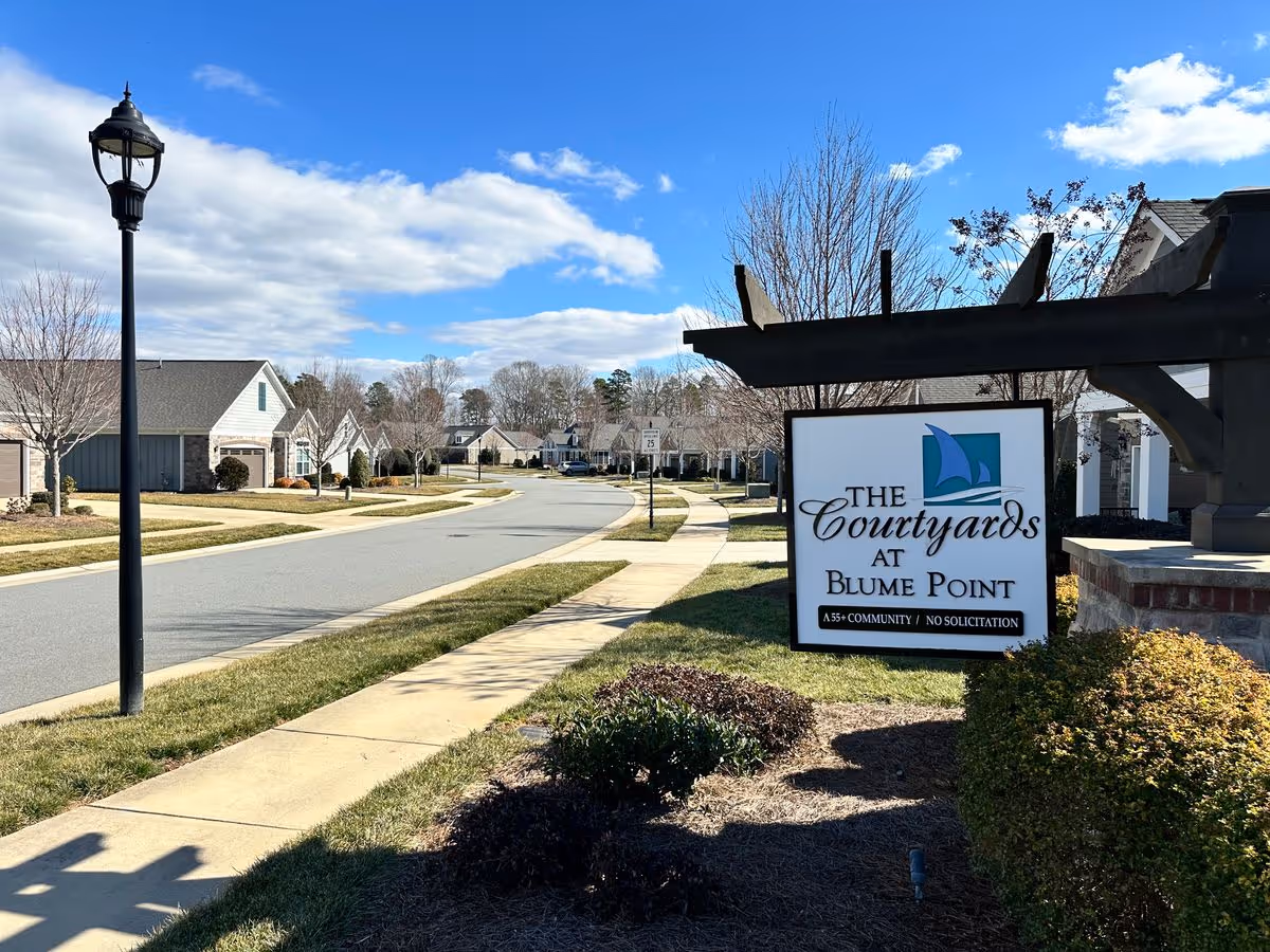 Neighborhood street with an entrance sign reading "The Courtyards at Blume Point" beside a sidewalk and single-story homes under a blue sky.