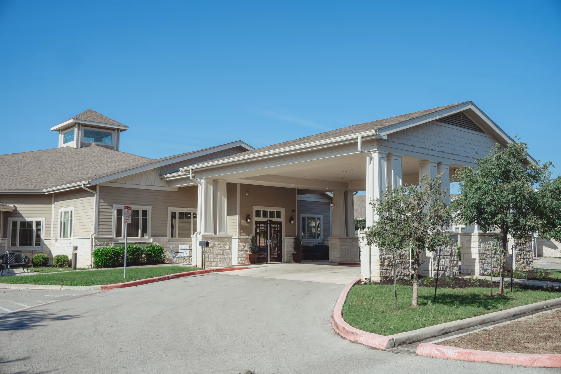 Front entrance of a single-story senior living facility with a covered porte-cochère, columns, and small landscaped areas under a clear blue sky.