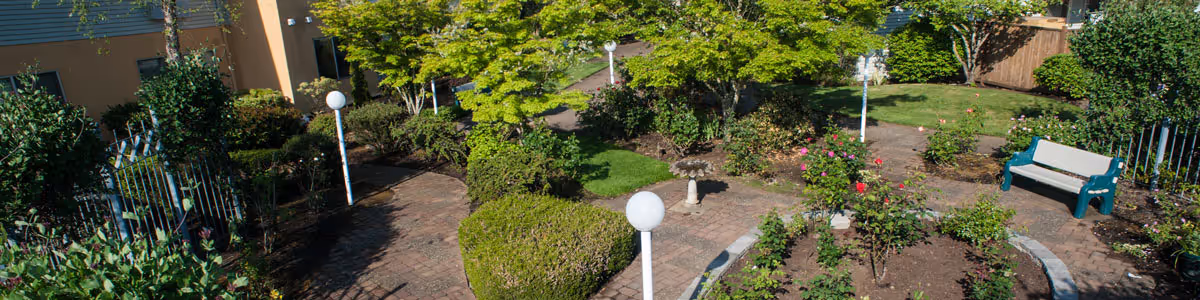 Small landscaped courtyard with brick paths, trimmed shrubs and flowerbeds, white globe lamp posts and a teal bench.