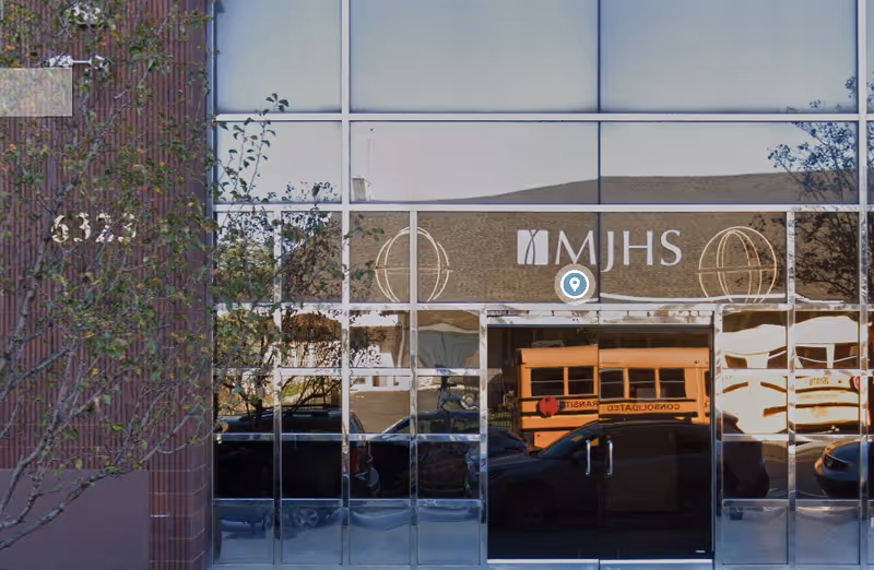 Front of a building with a glass facade reflecting vehicles and an MJHS sign.