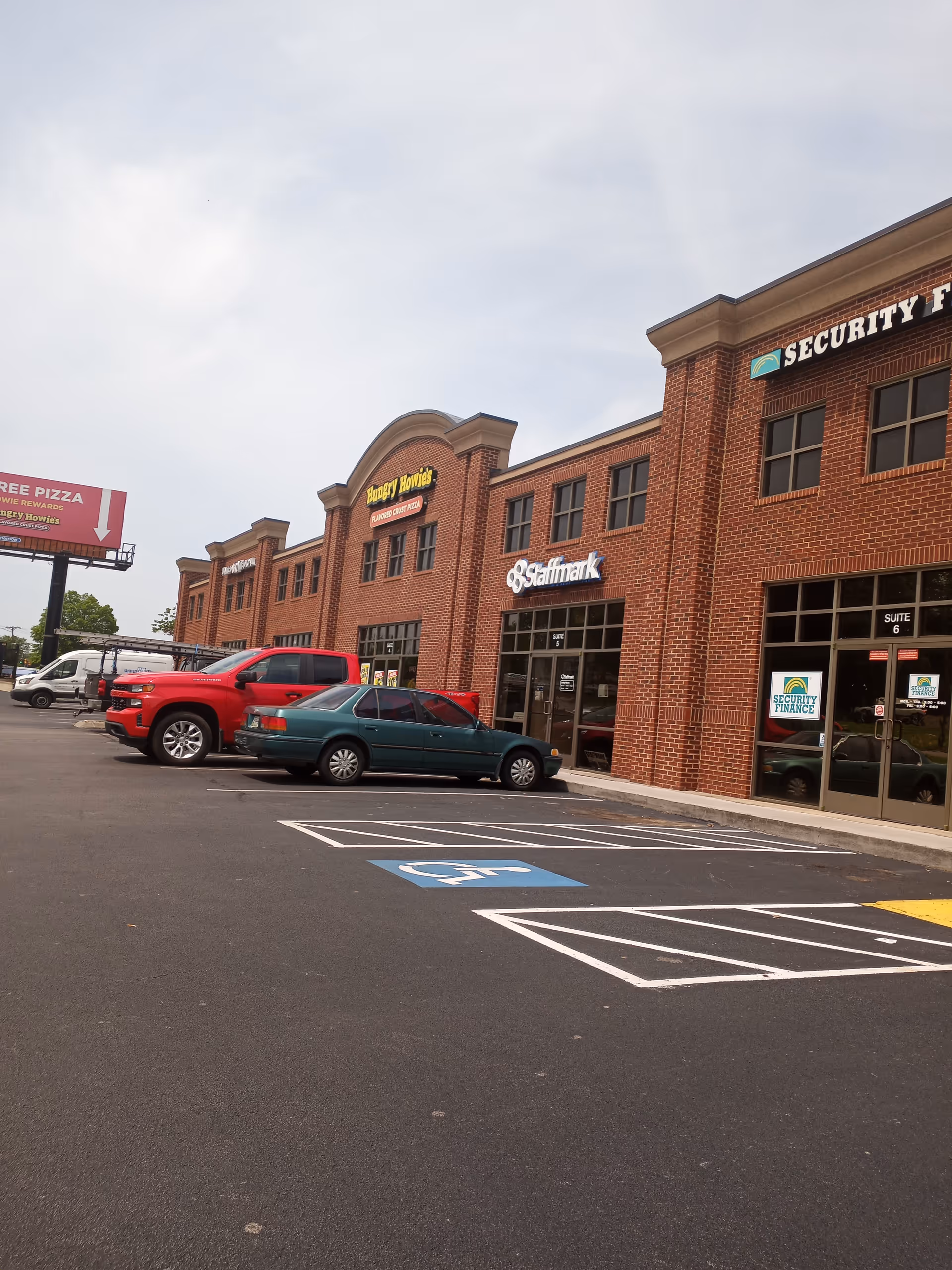 Brick storefronts line a parking lot with several cars and accessible parking.
