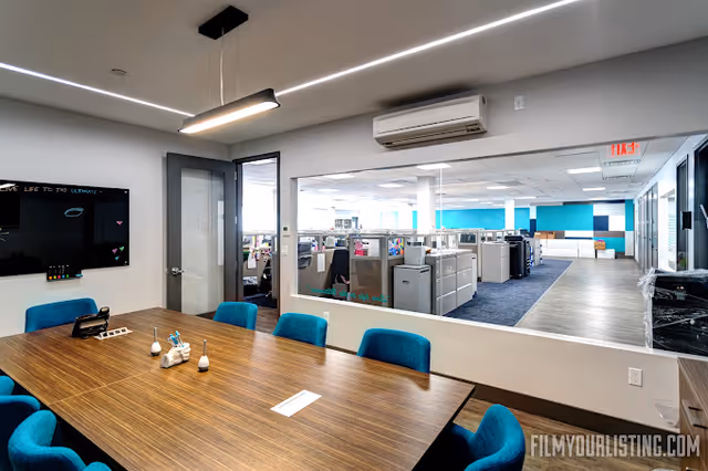 Conference room with wooden table, blue chairs, and glass wall revealing open office area.