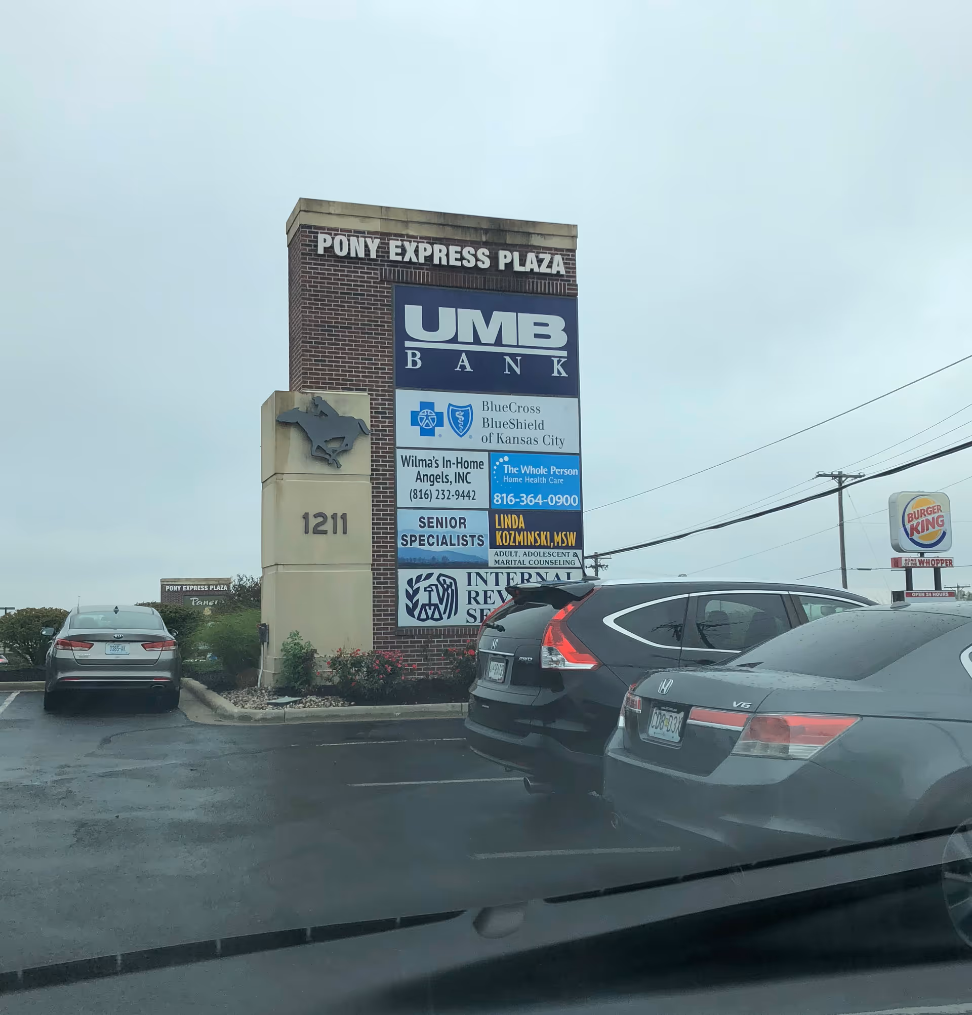 Tall brick plaza sign displays multiple business advertisements with parked cars outside.