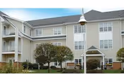 Multi-story beige senior living building with balconies, entryway, and landscaped lawn.