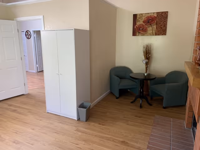 A cozy corner in a living area featuring two green armchairs around a small round black table with a decorative vase and lamp. A floral painting hangs on the beige wall above the chairs. To the left, there is a white cabinet and an open door leading to another room with wooden flooring. The floor is light wood, and part of a brick fireplace is visible on the right.