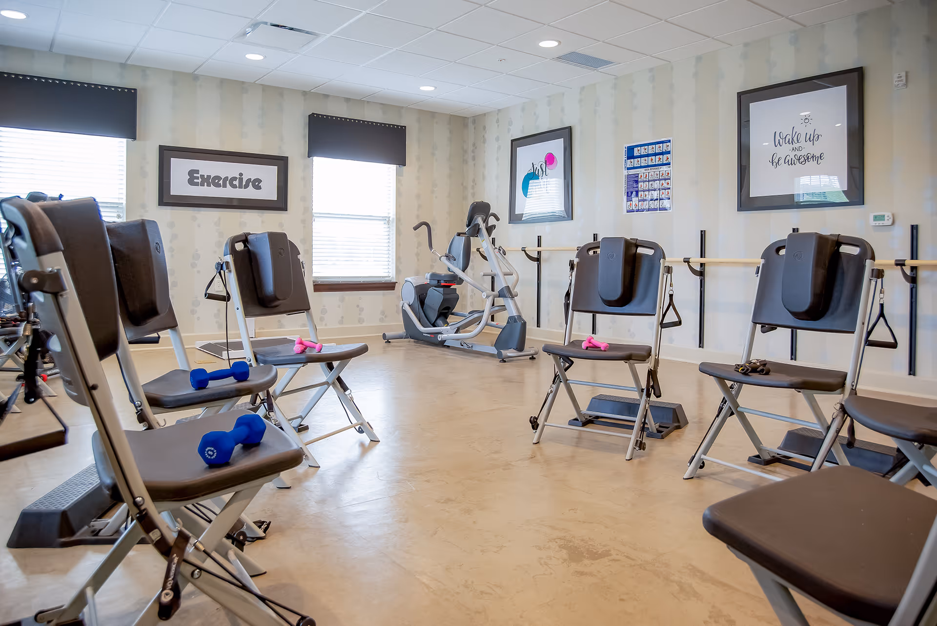 Senior living exercise room with chairs, cardio machines, weights, and wall art.