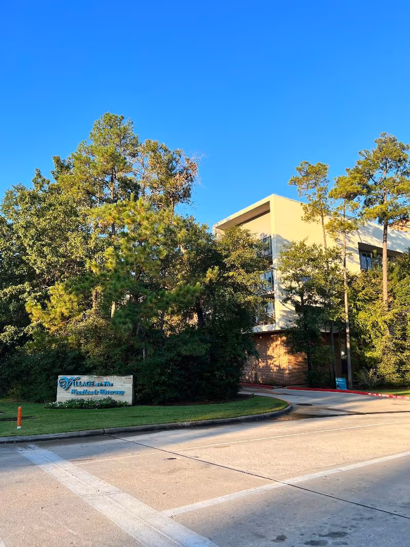 Exterior view of a senior living facility named Village at Woodlands Waterway, showing a multi-story building partially obscured by tall trees and greenery under a clear blue sky. A sign with the facility's name is visible near the entrance driveway.