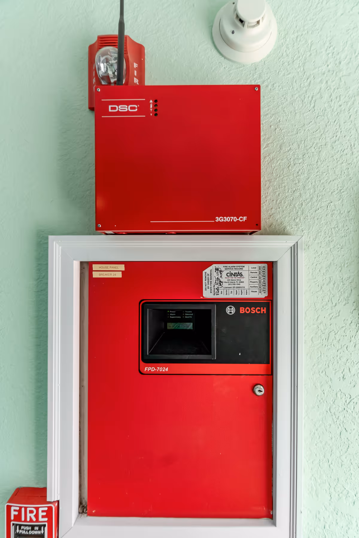 Close-up view of a red fire alarm control panel mounted on a light green textured wall, featuring a DSC unit on top, a Bosch panel below, a fire alarm pull station on the left, and a smoke detector on the upper right.
