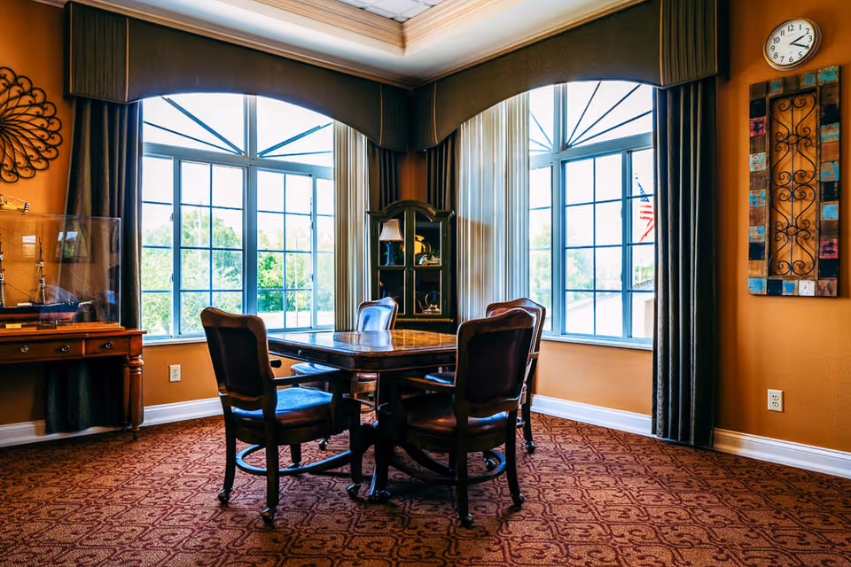 A cozy room with a square wooden table surrounded by four wooden chairs with dark cushions. The room has large windows with decorative curtains allowing natural light to fill the space. There is a wooden display cabinet in the corner and wall decorations including a colorful framed piece and a metal wall art. The carpet has a patterned design in warm tones.