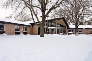 Snow-covered, single-story brick senior living building with glass entrance and leafless trees.
