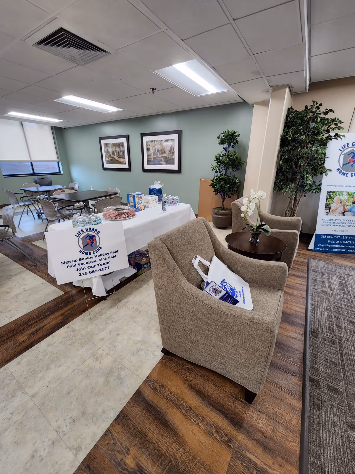 Indoor common area with tables, chairs, plants, framed art, and a Life Guard Home Care banner.