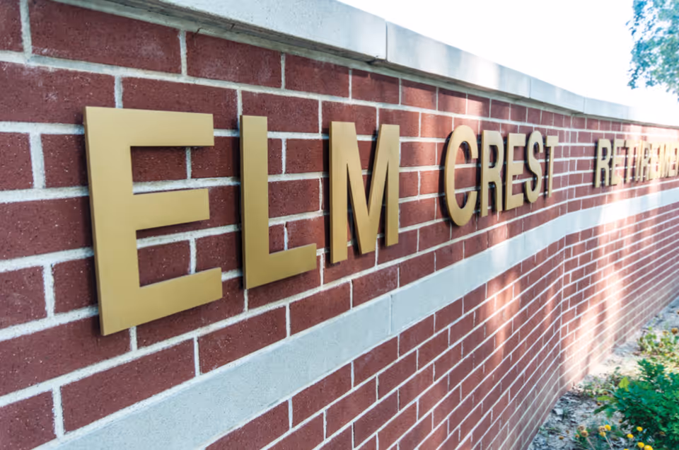 Close-up view of a red brick wall with large gold letters spelling out 'ELM CREST RETIRING' with some greenery visible at the base of the wall and trees in the background.