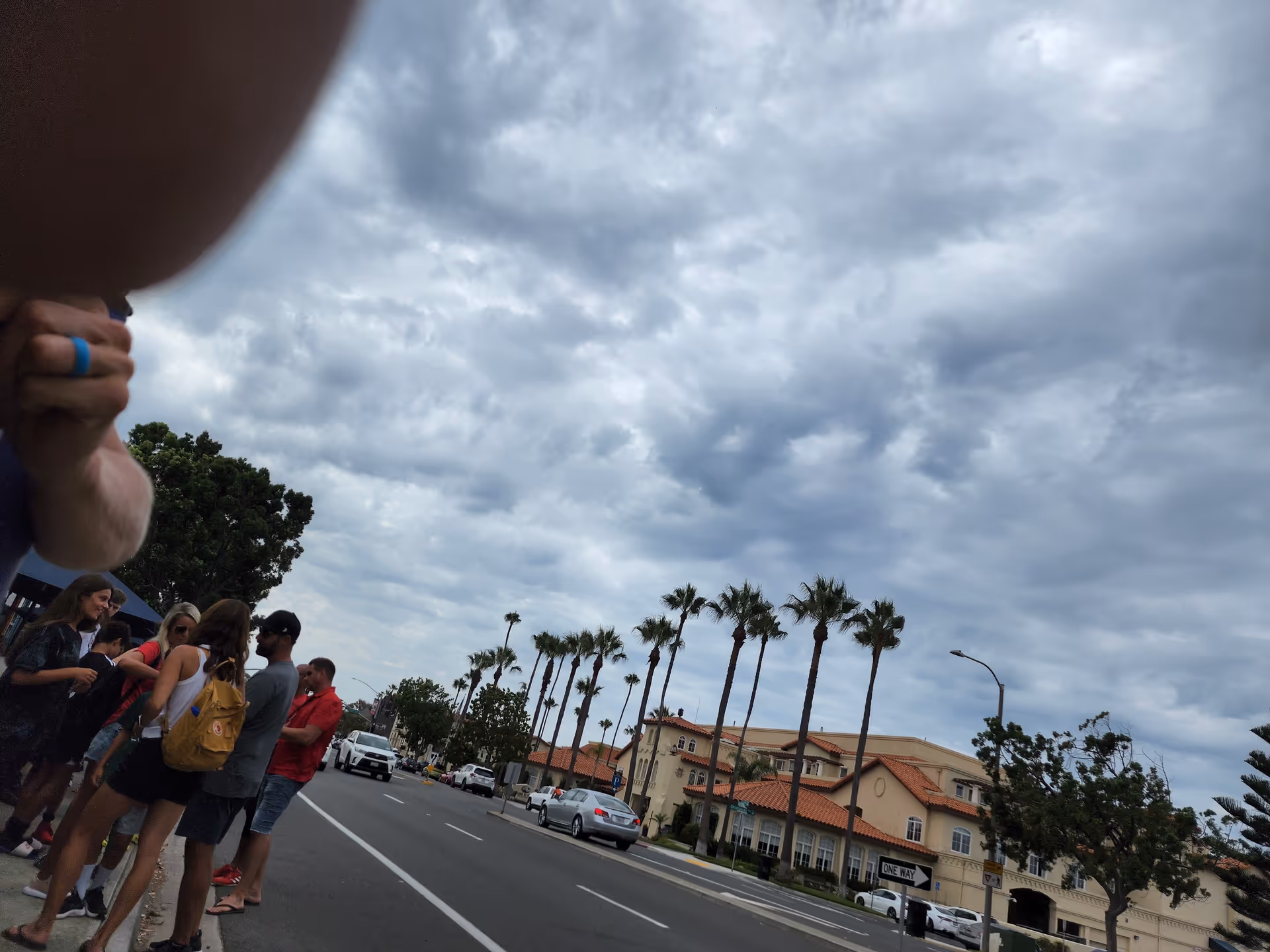 People gather on a sidewalk beside a tan building with palm trees along a street.