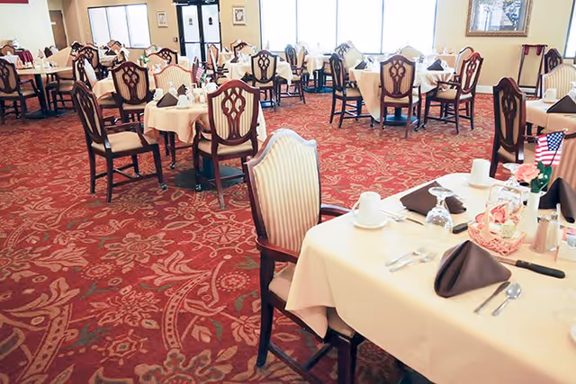 Spacious dining room with multiple round and rectangular tables set with place settings and chairs on a red patterned carpet.