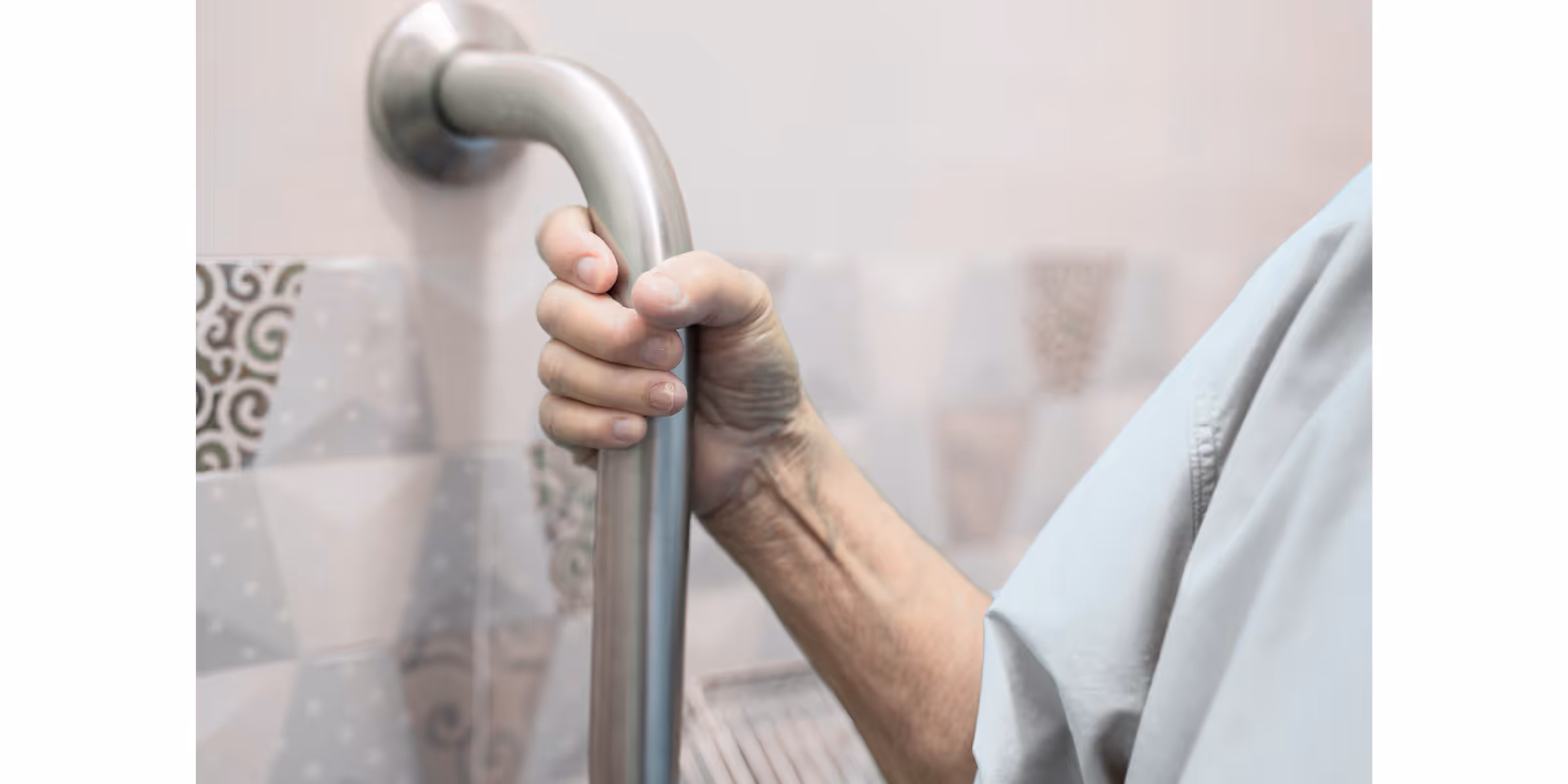 Close up of an elderly hand gripping a metal bathroom grab bar beside tiled wall.