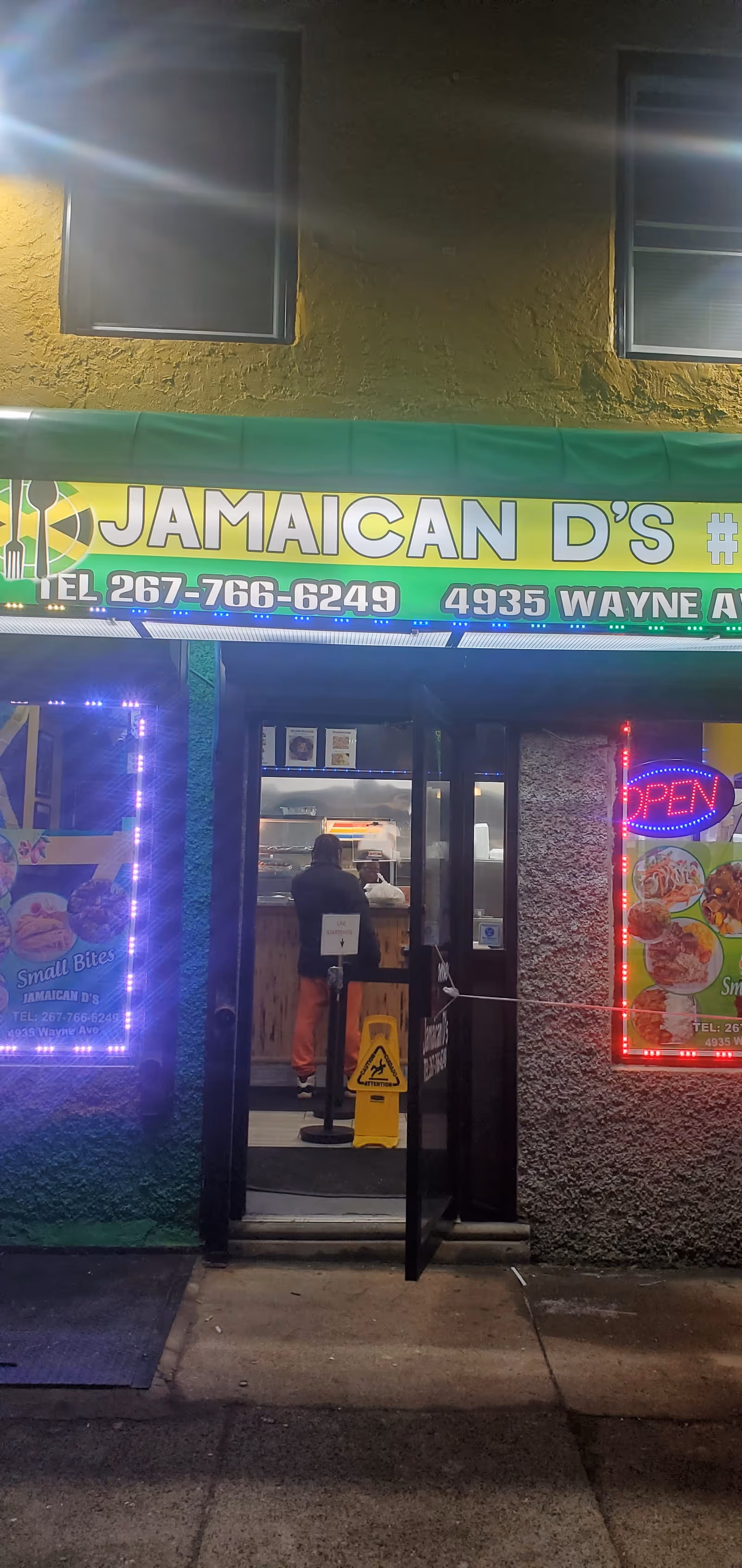 Nighttime storefront of Jamaican D's restaurant with open door and neon lights.