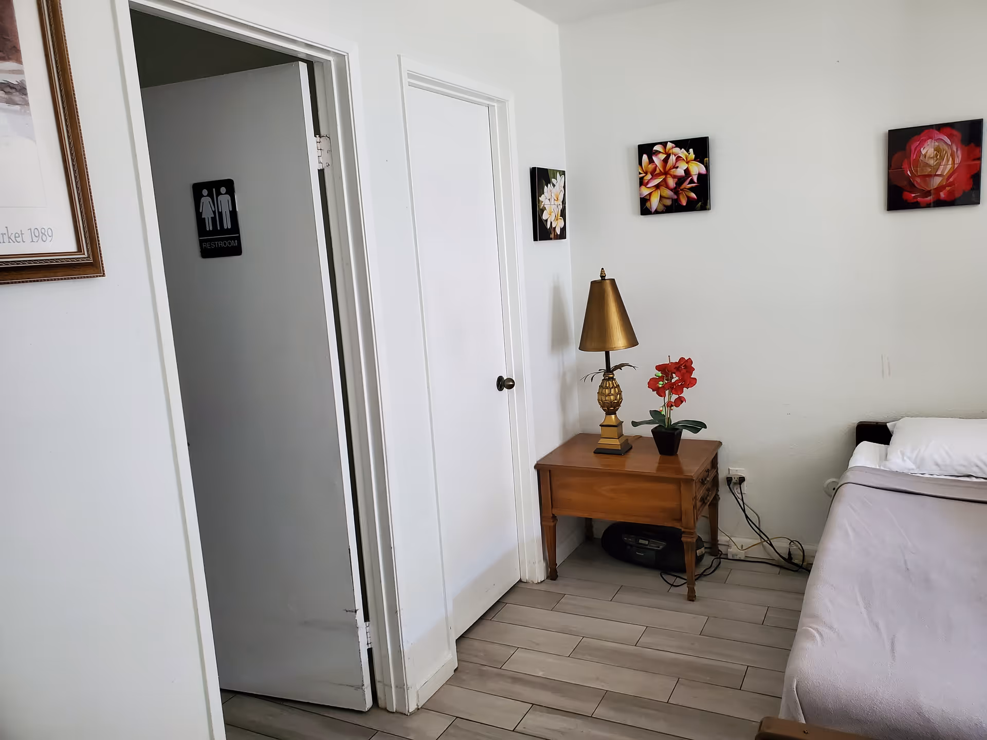 Bedroom interior with bed, nightstand, lamp, and floral wall art beside a restroom door.