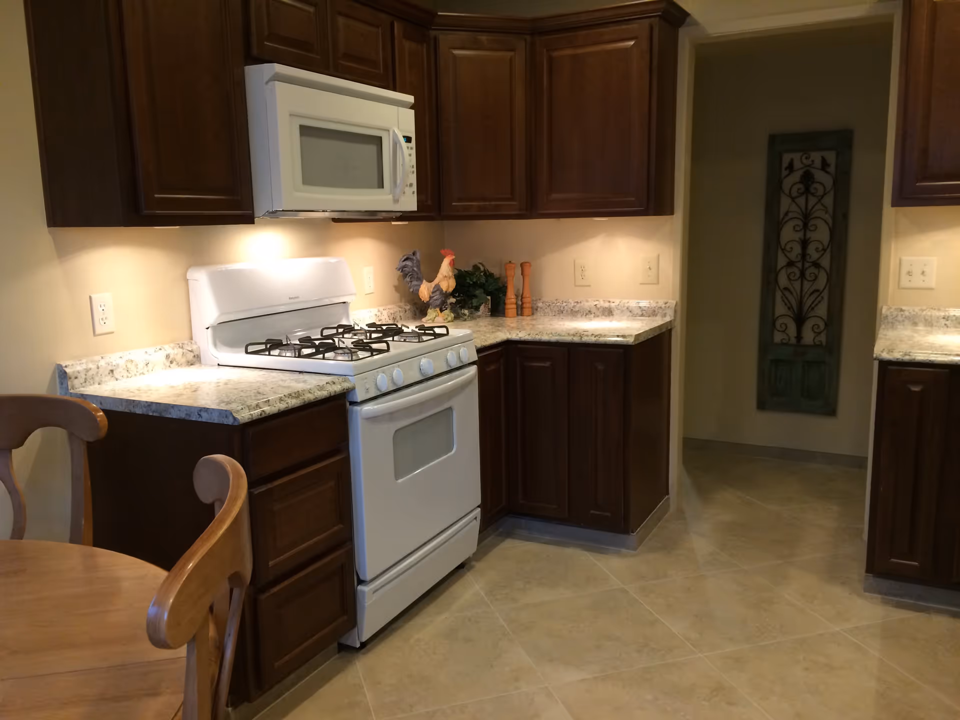 A kitchen with dark wooden cabinets, a white microwave mounted above a white gas stove, granite countertops, and a wooden dining table with chairs. There is a decorative rooster and salt and pepper shakers on the countertop, and a decorative wall hanging is visible in the hallway.