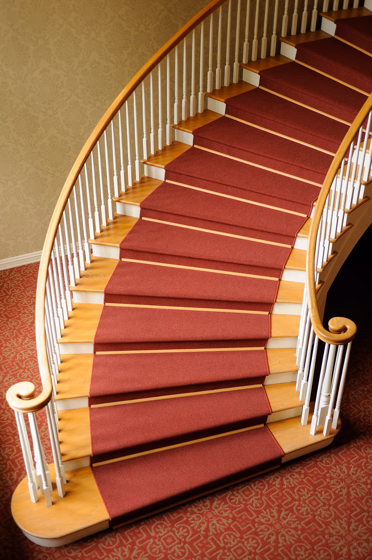 Curved wooden staircase with red carpet runner and white railings inside an elegant interior.