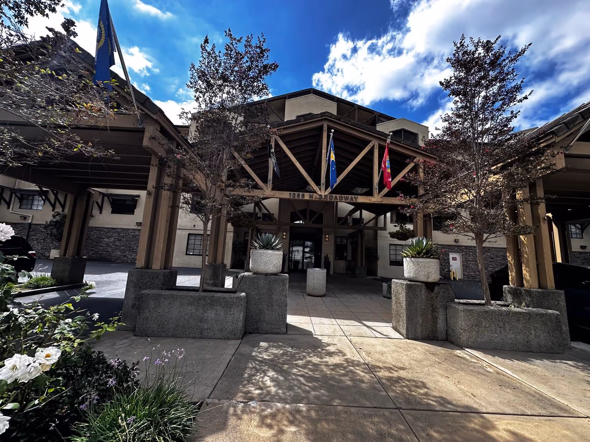 Front entrance of a senior living building with a covered porte-cochere, flags, planters, and trees under a partly cloudy sky.
