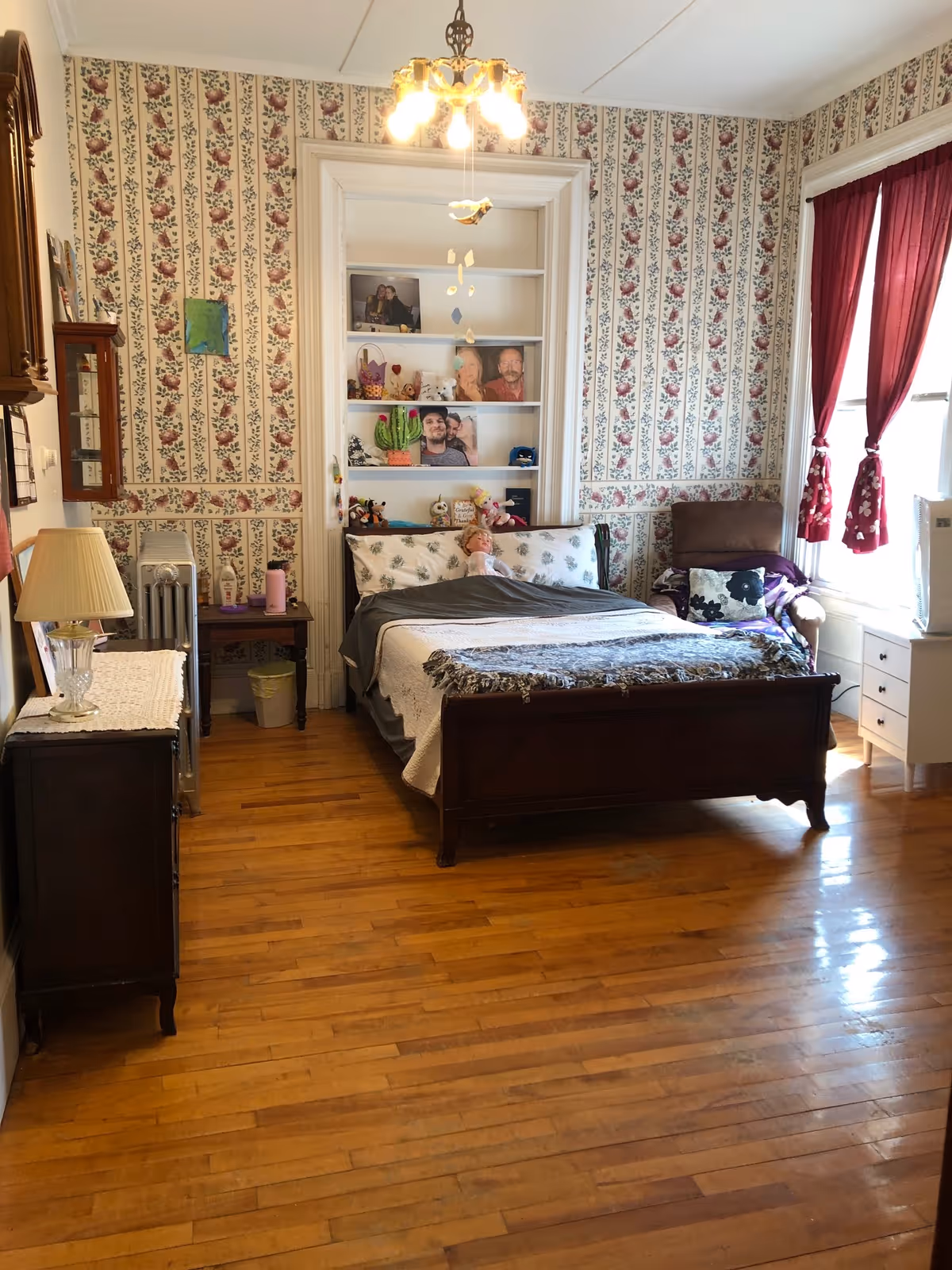 A cozy bedroom with floral wallpaper, a wooden bed with white and gray bedding, a doll sitting on the bed, a wooden dresser with a lamp, a small side table with a pink water bottle, a cushioned armchair with pillows near a window with red curtains, and built-in shelves with photos and decorative items.